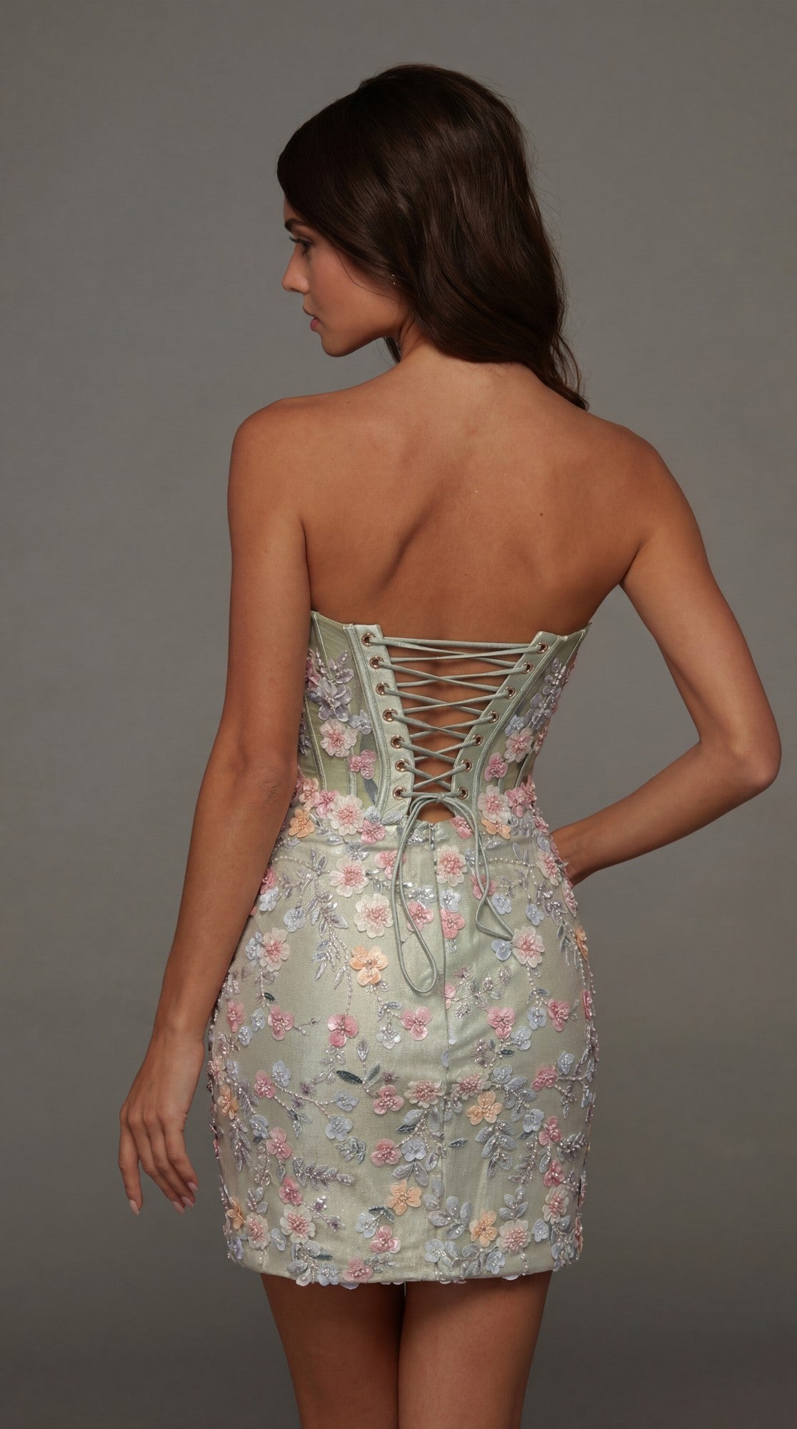 A woman stands facing away against a plain gray background, wearing the Alyce 40139 short homecoming dress with a strapless corset bodice, lace-up back, pastel tones, and delicate floral appliqués.